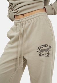Beige sweatpants met elastische tailleband en trekkoord, met een zwart "BROOKLYN NEW YORK" en een luipaardafbeelding op de dij.