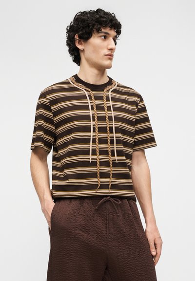 Homme aux cheveux bouclés portant un t-shirt rayé marron avec des cordons de serrage et un short marron texturé, les mains dans les poches, regardant vers la droite.