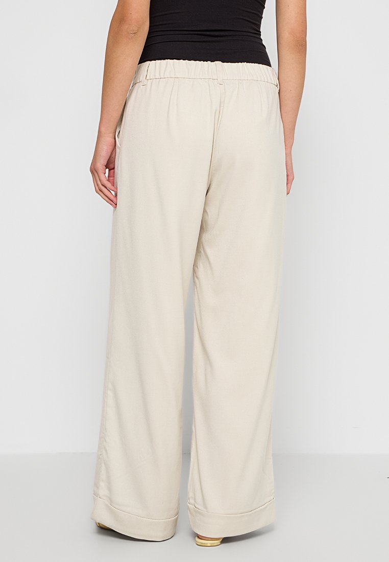 Beige weite Beinkleider mit elastischem Bund, glatter Textur und hochgekrempelten Säumen, von hinten vor einem neutralen Hintergrund dargestellt.