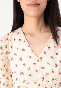 Blusa floral de tejido ligero con base color crema, que presenta pequeños patrones de rosas rojas, escote en V y acentos con mangas plisadas.
