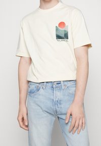 Camiseta de algodón crema con mangas cortas, que presenta un diseño gráfico en el bolsillo en verde y naranja que representa colinas y un sol, con la palabra "BALANCE".