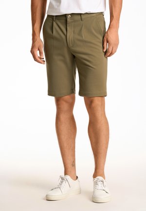Pantalones cortos de color verde oliva con frente plano, que presentan dos pliegues frontales, bolsillos laterales y un dobladillo recto, combinados con zapatillas blancas.
