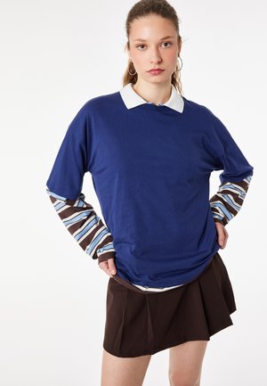 Ung kvinde med lys hud og håret sat op i en hestehale iført en blå kortærmet skjorte over en stribet langærmet trøje og et mørkt plisseret nederdel.