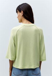 Light green, boxy top made from soft fabric, featuring three-quarter sleeves, a ribbed neckline, and a relaxed fit. Visible seams at shoulders.