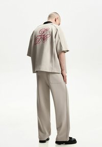 Beige overhemd met korte mouwen en een zwarte kraag, met een rood geborduurd logo op de achterkant. Loszittende broek met een gladde textuur, gedragen met zwarte schoenen.