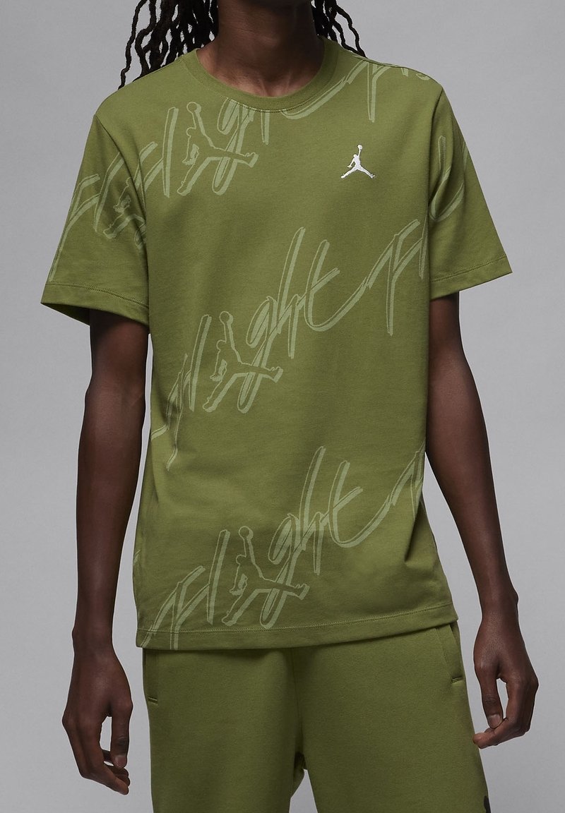 Personne portant une chemise à manches courtes vert olive et un pantalon avec un texte abstrait gris clair et un petit logo Jumpman blanc sur la poitrine.