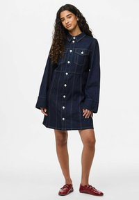 Femme aux longs cheveux bouclés, portant une robe en denim bleu foncé à boutons et des mocassins rouges, se tenant devant un fond blanc uniforme.