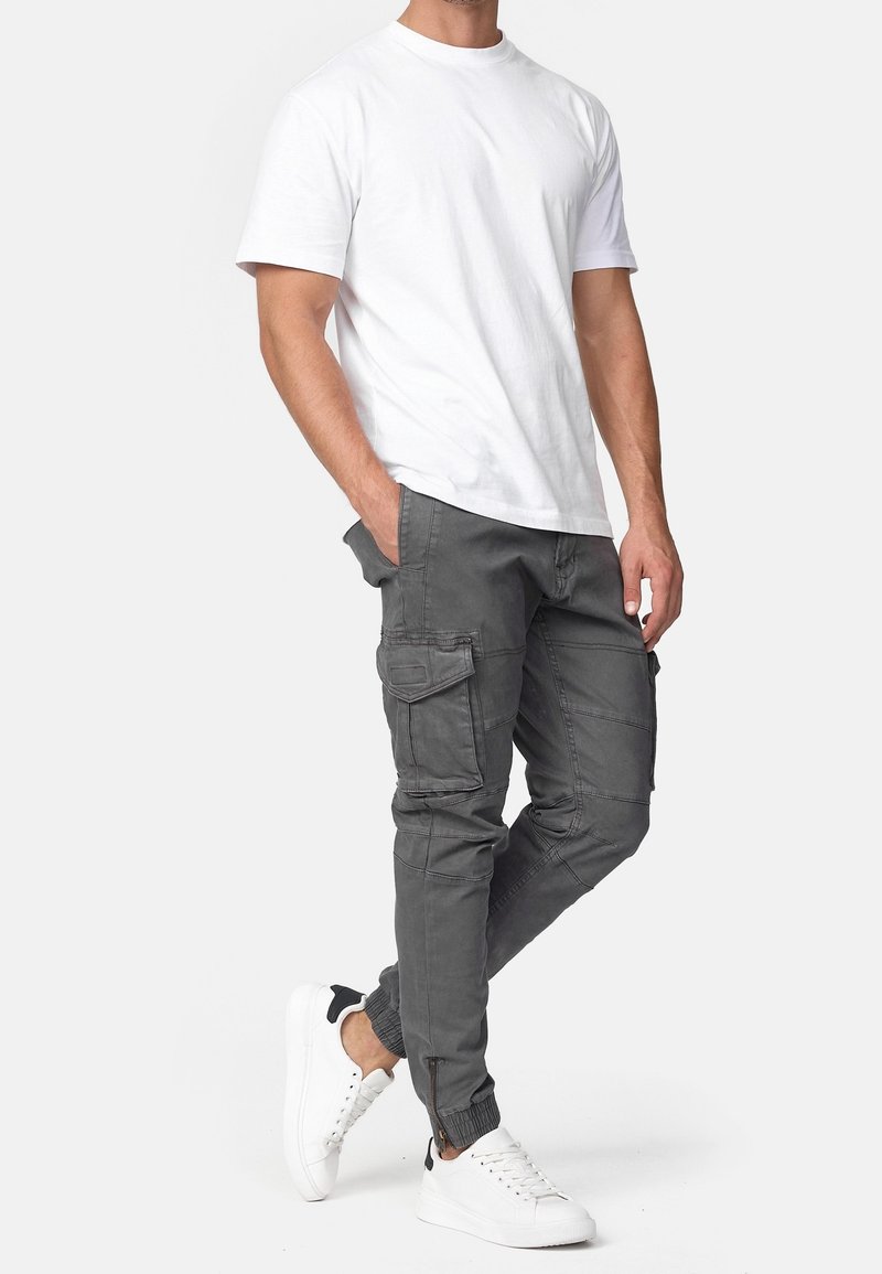 Homme portant un t-shirt blanc uni à manches courtes, un pantalon cargo gris avec plusieurs poches et des baskets blanches avec des détails noirs au talon, debout la main dans la poche.