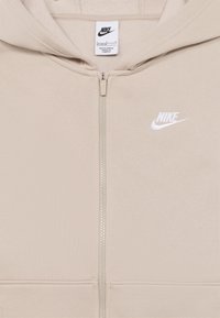 Μπεζ φούτερ με φερμουάρ κατασκευασμένο από μαλακό ύφασμα, με λευκό λογότυπο Nike στο αριστερό μέρος του στήθους και φερμουάρ στο ίδιο χρώμα, με κουκούλα και τσέπες μπροστά.