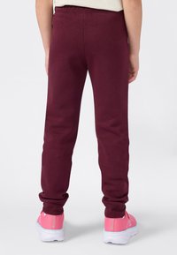 Barn som har på seg burgunder joggebukser og rosa sneakers, står med ryggen til på en enkel hvit bakgrunn.