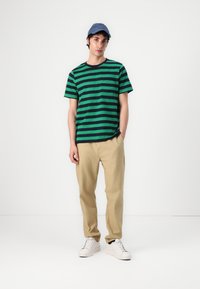 T-shirt rayé vert et marine avec un col rond, associé à un pantalon beige et des baskets blanches. Porté avec une casquette bleue.
