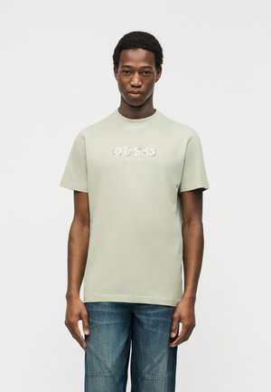 Jonge man staat recht vooruit kijkend, draagt een lichtgroen GUESS-logo T-shirt en blauwe spijkerbroek tegen een effen witte achtergrond.