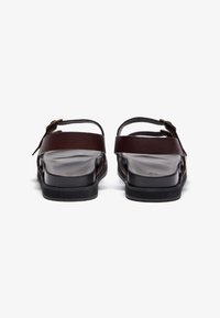 Brown leather sandals with a flat sole, adjustable ankle straps, and a smooth finish. Features a minimalist design and subtle hardware accents.
