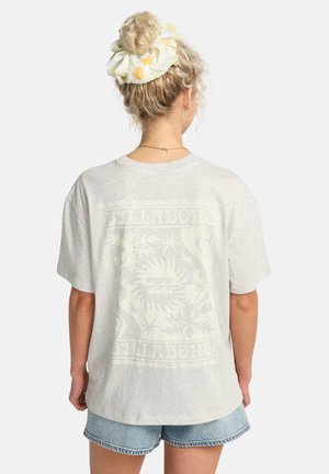 Donna con capelli biondi ricci raccolti in uno scrunchie floreale, indossa una t-shirt Billabong grigio chiaro e pantaloncini di jeans, vista da dietro.