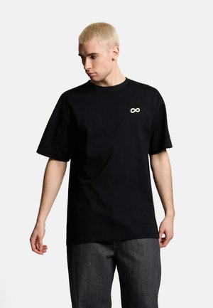 Blondyn noszący czarny, oversize T-shirt z białym symbolem nieskończoności na piersi i ciemnoszare jeansy, patrzący w dół w lewo.