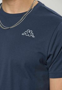 Άτομο που φοράει μπλε σκούρο T-shirt με λευκό κεντημένο λογότυπο δύο καθιστών φιγούρων πλάτη με πλάτη και ασημένια αλυσίδα λαιμού.