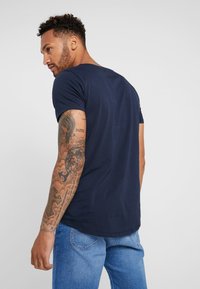 T-shirt in cotone blu navy con maniche corte e orlo arrotondato. Presenta un design aderente e tatuaggi visibili sul braccio.