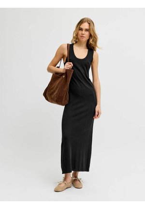 Femme portant une robe maxi noire sans manches, des ballerines beiges avec des nœuds, tenant un grand sac à bandoulière en daim marron sur un fond uni.