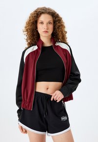 Młoda kobieta z kręconymi włosami, ubrana w burgundową i czarną kurtkę zapinaną na zamek, czarny crop top oraz czarne spodenki sportowe z białymi wykończeniami.
