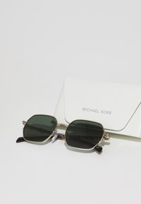 Hexagonale solbriller med mørkegrønne linser, gyldent metallisk ramme og sorte stangene, ledsaget av en hvit Michael Kors-etui.