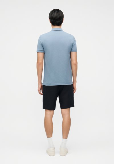 Άνδρας που στέκεται με την πλάτη γυρισμένη, φορώντας ανοιχτό μπλε πόλο πουκάμισο, μαύρες βερμούδες, λευκές κάλτσες και λευκά αθλητικά παπούτσια σε απλό φόντο.