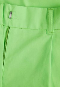 Dettaglio di tessuto verde brillante di pantaloni con cerniera e accento hardware rettangolare in argento in vita, con cuciture precise visibili.