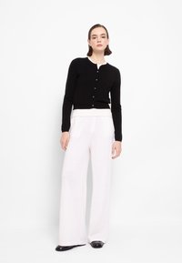 Cardigan nero con scollatura rotonda e bottoni, abbinato a pantaloni ampi rosa chiaro. Tessuto morbido e polsini aderenti, completati da scarpe nere.