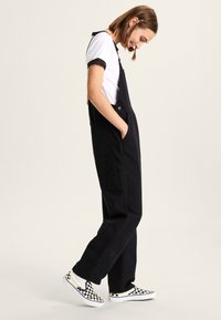 Zwarte overallbroeken met zijzakken, gedragen over een zwart-witte kraagshirt. Geprinte geruite instappers complementeren de outfit.