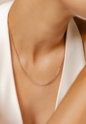 Thin silver chain necklace with a delicate design, resting on smooth skin, complements a white garment in the background.