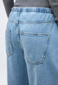 Κοντινό πλάνο ανοιχτό μπλε τζιν με ελαστική μέση και πίσω τσέπες, φορεμένο με γκρι ριγωτό πουλόβερ.