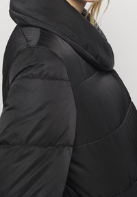 Chaqueta negra acolchada con una capucha grande y suave que se pliega, diseño acolchado, textura lisa y líneas de costura prominentes en el cuerpo.