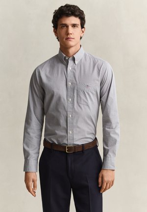 Jeune homme portant une petite chemise à carreaux boutonnée, rentrée dans un pantalon sombre avec une ceinture marron, se tenant devant un fond uni.