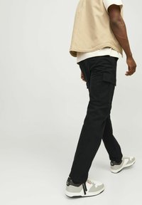 Pantaloni cargo negri cu buzunare laterale, asortați cu pantofi sport gri și albi. Ținuta include o jachetă bej fără mâneci deasupra unei cămăși deschise la culoare.