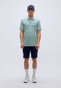 Ανοιχτό πράσινο πόλο μπλουζάκι με υφασμένο μοτίβο διαμαντιού, συνδυασμένο με σκούρα μπλε σορτς και λευκές κάλτσες. Τα λευκά αθλητικά παπούτσια ολοκληρώνουν την εμφάνιση.