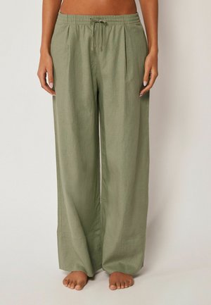 Kvinde iført olivengrønne bukser med vide ben, elastisk talje og snøre, stående barfodet mod en ensfarvet baggrund.