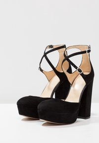 Černé semišové vysoké podpatky na platformě s kulatou špičkou a vysokým blokovým podpatkem. Obsahují zkřížené kotníkové popruhy s nastavitelnými sponami.