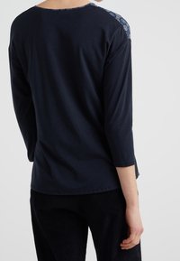 Tummansininen, pitkähihainen paita, jossa on pyöreä kaula-aukko; olkapäillä kukkakuviolliset yksityiskohdat; pehmeä, kevyt kangas; rento malli.