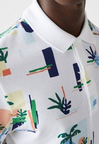 Polo à manches courtes en coton blanc avec des motifs abstraits colorés, comprenant des plantes et des formes géométriques, avec un col classique et des boutons.