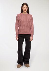 Różowy sweter z dzianiny w warkocze z długimi rękawami, okrągłym dekoltem, w połączeniu z czarnymi spodniami o szerokich nogawkach i brązowym obuwiem wsuwanym.