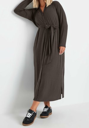 Kvinde iført en lang mørkebrunt ribbet kjole med knapper og et matchende bælte, kombineret med sorte og hvide afslappede sneakers.