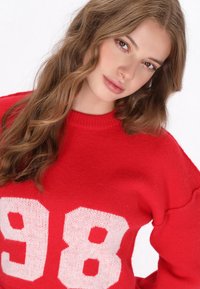 Roter gestrickter Pullover mit lockerer Passform, der an der Vorderseite große weiße Zahlen "9" und "8" zeigt. Weicher Stoff und runder Ausschnitt.