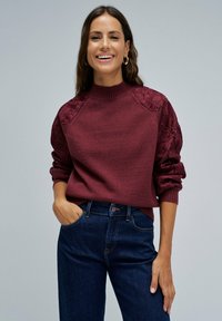Mujer sonriente vestida con un suéter burdeos con mangas de encaje floral y jeans de color azul oscuro, de pie con una mano en el bolsillo contra un fondo gris.