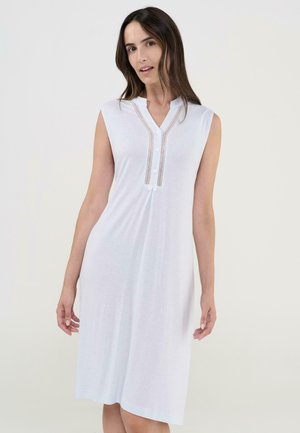 Mujer con vestido blanco sin mangas, de largo hasta la rodilla, con cuello en V abotonado y ribete de encaje, de pie frente a un fondo claro y liso.