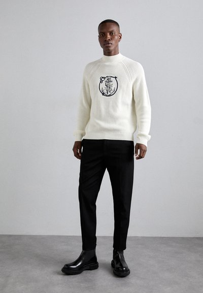 Suéter blanco acanalado con diseño bordado en azul marino, combinado con pantalones negros y botas de tobillo negras brillantes. Entorno minimalista con fondo gris.