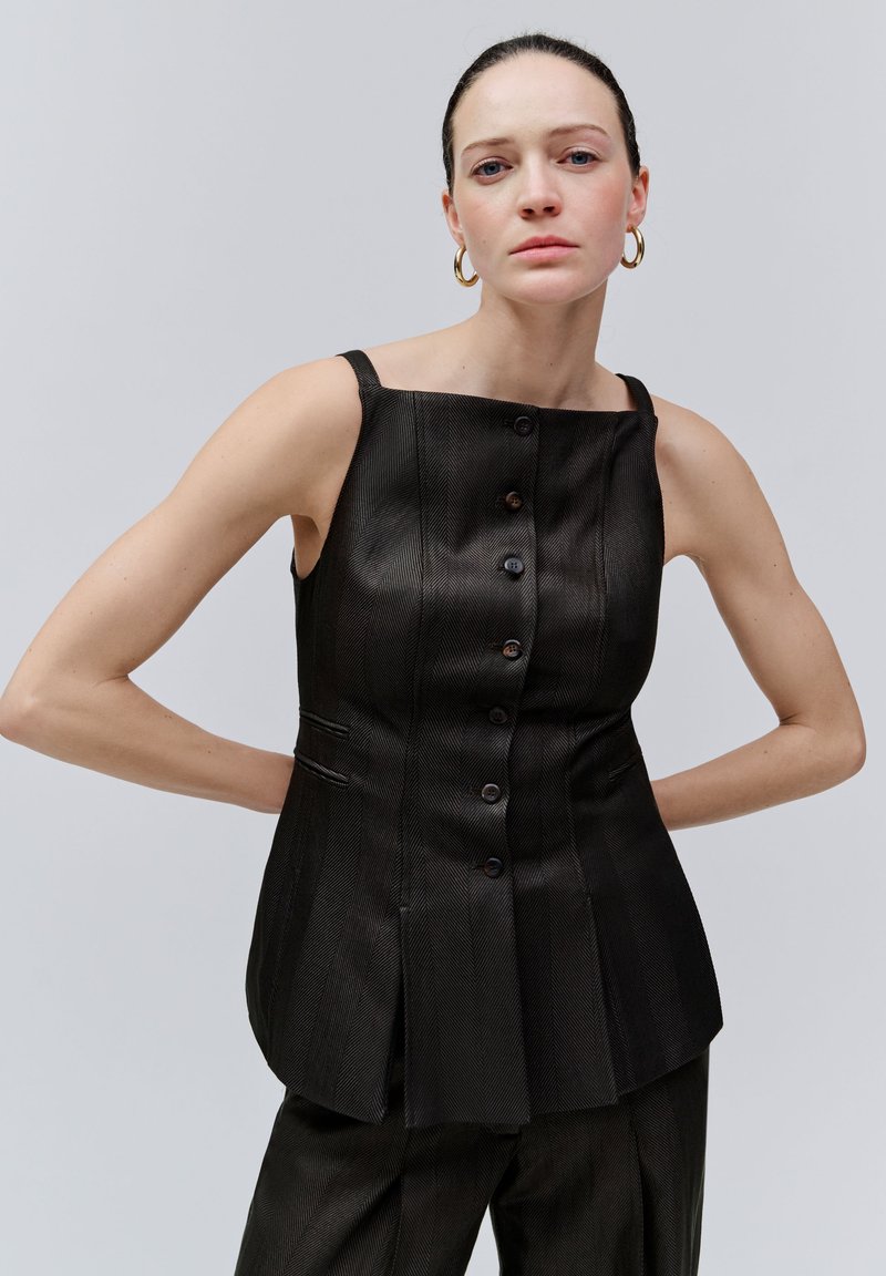 Jonge vrouw met donkere mouwloze top met knopen en bijpassende broek, staand met haar handen achter haar rug, neutrale grijze achtergrond.