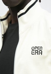 Hvid lynlåstrøje med sorte detaljer og ribbet tekstur, med "OPEN ERA" broderet på venstre brystområde.