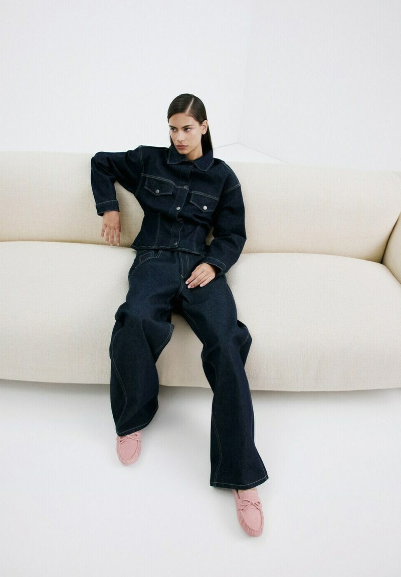 Kvinde iført mørk denimjakke og vidde jeans, der sidder på en beige sofa med den ene arm hvilede på ryggen.