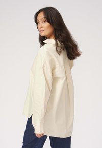 Lys beige oversized skjorte lavet af bomuldsstof, med spids krave, lange ærmer og sideslidser ved kanten. Glat tekstur og afslappet pasform.