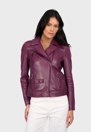 Vrouw met lang donker haar, gekleed in een paarse leren motorjack over een bruine coltrui en witte broek, staand tegen een effen achtergrond.