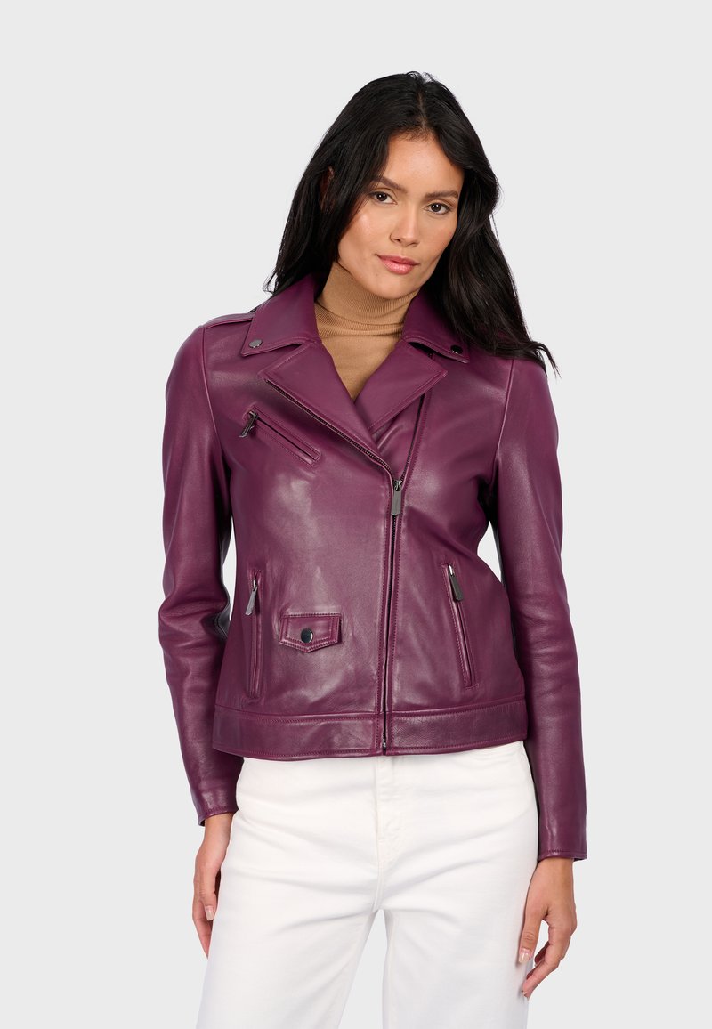 Žena s dlouhými tmavými vlasy, oblečená v purpurové kožené motorkářské bundě přes hnědý rolák a bílé kalhoty, stojící proti jednobarevnému pozadí.